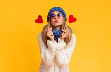 Lovely girl in winter hat and sunglasses holding red heart cards and showing kissing lips, pink background