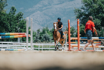 Equestrian rider training on horseback over colorful jumps in an outdoor riding arena with a trainer observing.