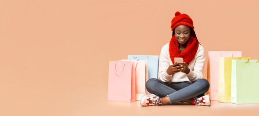 A cheerful young black woman wearing a red hat and scarf sits on the floor, happily using her smartphone. Colorful shopping bags are arranged around her, copy space