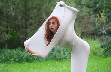 A woman stretches a translucent fabric around her body in a serene outdoor setting. Her expression...