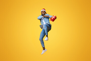Christmas delivery concept. Positive african american guy jumping with Xmas present, hurry to deliver gift box in time. yellow background