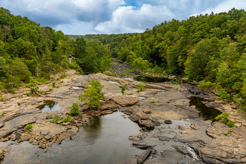 A summer view of the top of Little River Falls in Little River Canyon National Preserve, Alabama. Lush green foliage lines the river bed, and shallow pools of water form in the rock layers.