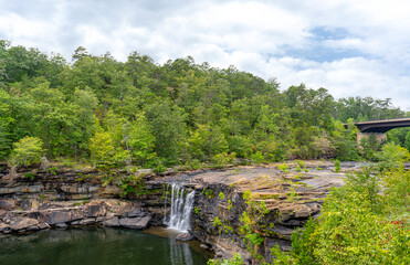 Fototapeta premium In the summer, Little River Falls in Little River Canyon National Preserve in Alabama is surrounded by green forests set against a cloudy blue sky. The Hwy 35 bridge is visible in the background.