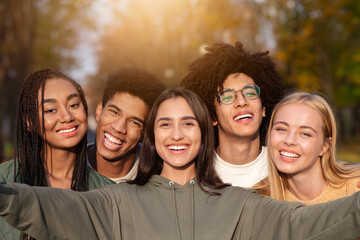 Smiling teen international friends taking selfie while walking in autumn park, happy memories concept