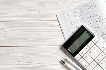 Copybook with handwritten maths formulas, calculator and stationery on white wooden background