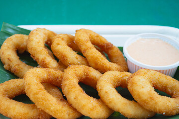 Golden onion rings served with sauce in biodegradable tray on green leaf.