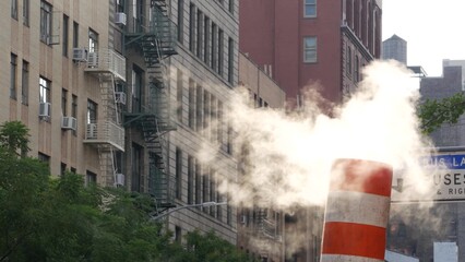 Steam vapor vented on New York City street, orange vapour tube stack. Hot smoke from pipe. Con Edison's Steam Operations system, Manhattan. Morning light, fog. Only bus lane. Rooftop water tower tank.