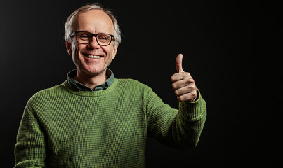 Naklejka premium Cheerful adult male in a green sweater and glasses with a thumbs-up gesture and a broad smile, set against a black background. Portrait capturing confidence and satisfaction.