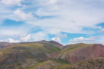 Beautiful layered landscape with multicolor hills and mountains in sunlight under clouds in blue sky. Scenic alpine view to sunlit colorful mountain range of many vivid colors in changeable weather.
