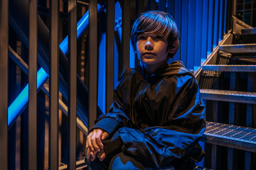 A young boy, dressed in a dark jacket, sits on metal steps at night, deep in thought. The...
