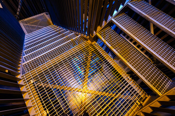 A detailed view of a metal staircase with grid-patterned steps, illuminated by blue and yellow...
