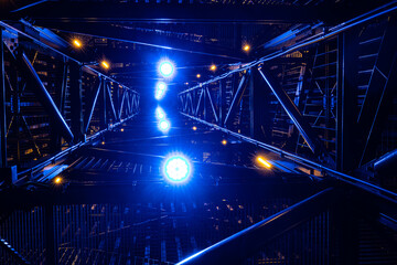 An upward view of a metal structure at night, dramatically lit with bright blue and yellow lights. The intricate framework and vibrant lighting create a futuristic, abstract visual effect.