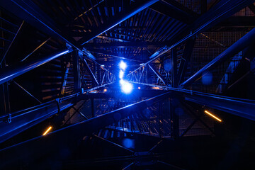 An upward view of a metal structure at night, dramatically lit with bright blue and yellow lights....