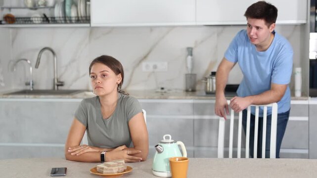 Sad young girl sitting at the table while her boyfriend is quarreling with her standing behind in the kitchen