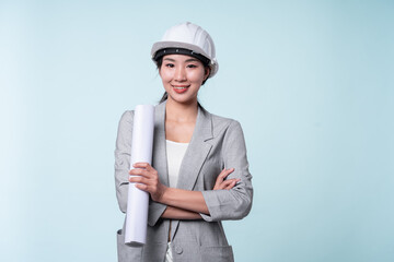 Young Asian female engineer or architect wearing white helmet holding blueprint isolated light blue background.