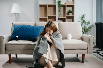 Obraz premium Young Asian woman suffering from flu symptoms, covered with a blanket on a sofa. Concept of illness, healthcare, and recovery.