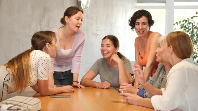 Smiling friendly woman conducting informal work meeting with mixed age group of female colleagues sitting on chairs in circle in office, discussing plans, projects, or work-related situations. High