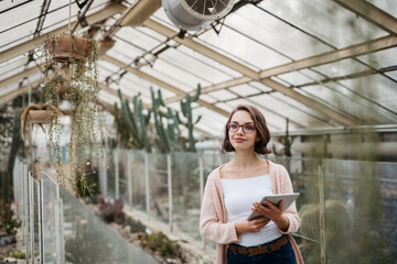 Small greenhouse business. Businesswoman selling flowers and seedlings, standing in greenhouse, looking at camera, smiling.