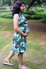 Fototapeta premium A pregnant Indian lady poses for outdoor pregnancy shoot and hands on belly, Indian pregnant woman puts her hand on her stomach with a maternity dress at society park, Pregnant outside maternity shoot