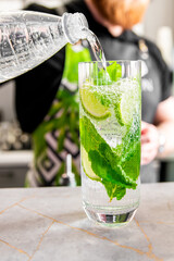 Close-up of a refreshing mojito cocktail with lime and mint being poured from a bottle. Vibrant green colors of mint and lime against the clear liquid suggest freshness, ideal for culinary themes.