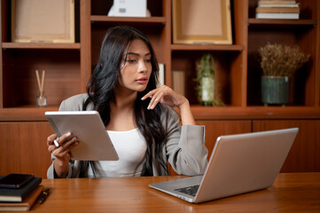 A businesswoman is focusing on her work, holding a tablet while reading online papers on laptop.