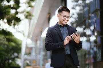 Confident Asian businessman in modern city, smiling while using smartphone outdoors, wearing professional suit, representing success and connectivity in digital age.