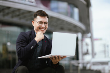Fototapeta premium A determined young businessman in a city, confidently working on his laptop outdoors, dressed in a suit, embodying leadership, success, and motivation in the corporate world.