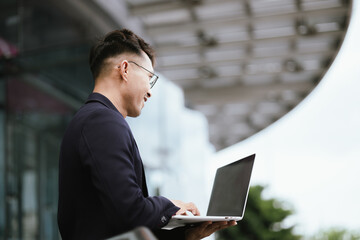 A determined young businessman in a city, confidently working on his laptop outdoors, dressed in a...