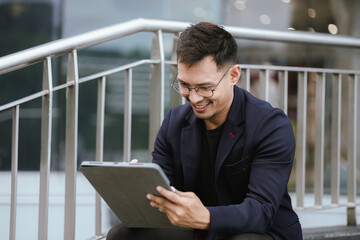 Fototapeta premium A cheerful young Asian businesswoman in a modern city, smiling confidently while using her smartphone outdoors, dressed in a professional suit, embodying success and digital connectivity.