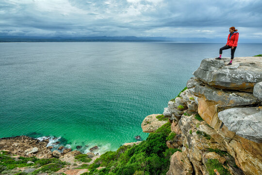 Woman standing on edge of cliff and looking at ocean in Robberg Nature Reserve, Garden Route National Park, Western Cape, South Africa