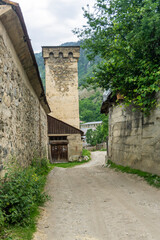 Medieval defensive stone tower with loopholes at the end of a street in a village. Stone walls on the sides. Earthen road, trees