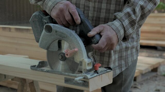 Close-up of an elderly man's hands with a circular saw. The man skillfully operates a circular saw to cut wood in an outdoor workshop.