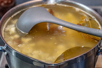 Delicious cooked soup on a meaty clear broth of smoked pork beef ribs with potatoes, cauliflower, bay leaf and salt in a stainless steel pot close-up with a plastic gray ladle.