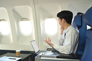Fototapeta premium Side view of asian male passenger talking on his cellphone while sitting in an airplane seat with laptop