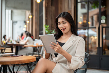 A beautiful, happy Asian businesswoman is using her digital tablet, working remotely at a cafe.