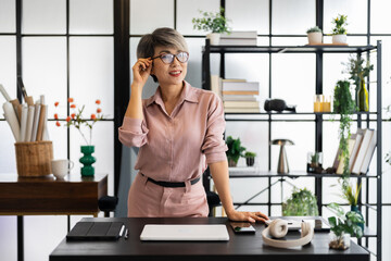 aged businesswoman with short gray hair stands confidently at her desk, hands resting on the table,...
