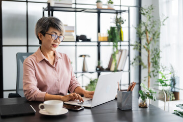 A middle-aged businesswoman with short gray hair is working on her laptop in a modern office, surrounded by a stylish, green-filled environment