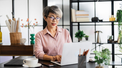 A middle-aged businesswoman with short gray hair is working on her laptop in a modern office, surrounded by a stylish, green-filled environment