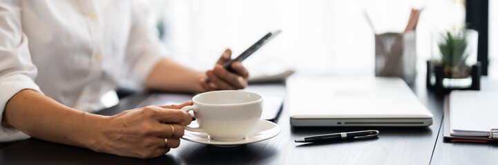 A middle-aged businesswoman is seated at her desk, holding a smartphone and working on it in a modern, stylish office setting