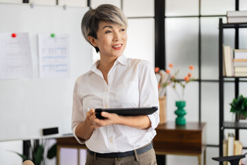 A female architect stands thoughtfully, holding a tablet in a modern office filled with design books and materials, ready to present her ideas