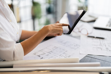 A female architect reviews architectural plans on her tablet while sitting at her desk, with various design sketches and tools around her in a modern, well-organized office