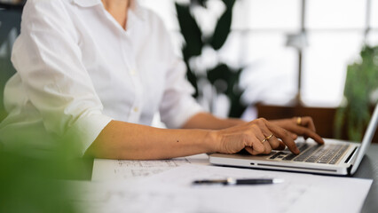 A female architect works on her laptop, surrounded by blueprints and architectural drawings, concentrating on her design projects in a tidy and modern office setting