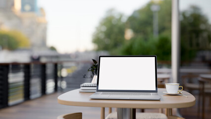 A laptop computer with a white-screen mockup on a table on a coffee shop outdoor terrace or balcony.