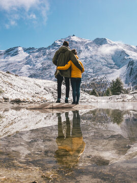 two persons holding each other in green and yellow jacket reflection in a calm pond in the mountains with snowy mountains in distance