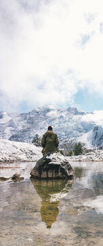 person in green jacket reflection in a calm pond in the mountains with snowy mountains in distance
