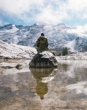 person in green jacket reflection in a calm pond in the mountains with snowy mountains in distance
