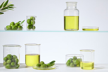 Several glassware displayed on two tier lab shelf, filled by olive oil and fresh olive, which is popular ingredient in moisturizers, serums, and face masks. Front view, displaying space