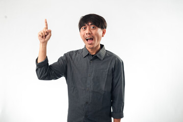 Fototapeta premium An enthusiastic Asian man in a gray shirt pointing upward with one hand while the other hand is on his hip. He has a wide, excited expression, standing against a plain white background.