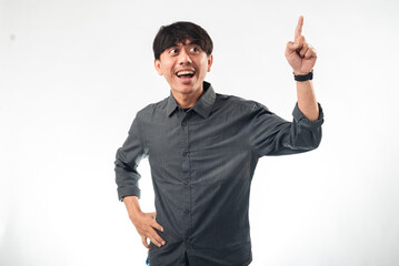 Fototapeta premium An enthusiastic Asian man in a gray shirt pointing upward with one hand while the other hand is on his hip. He has a wide, excited expression, standing against a plain white background.