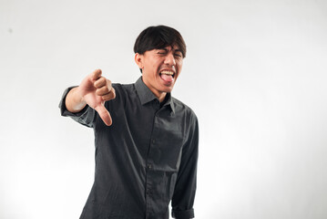 Naklejka premium An Asian man in a gray shirt making a disapproving face while giving a thumbs-down gesture. He is sticking out his tongue, expressing dislike or disappointment, against a plain white background.